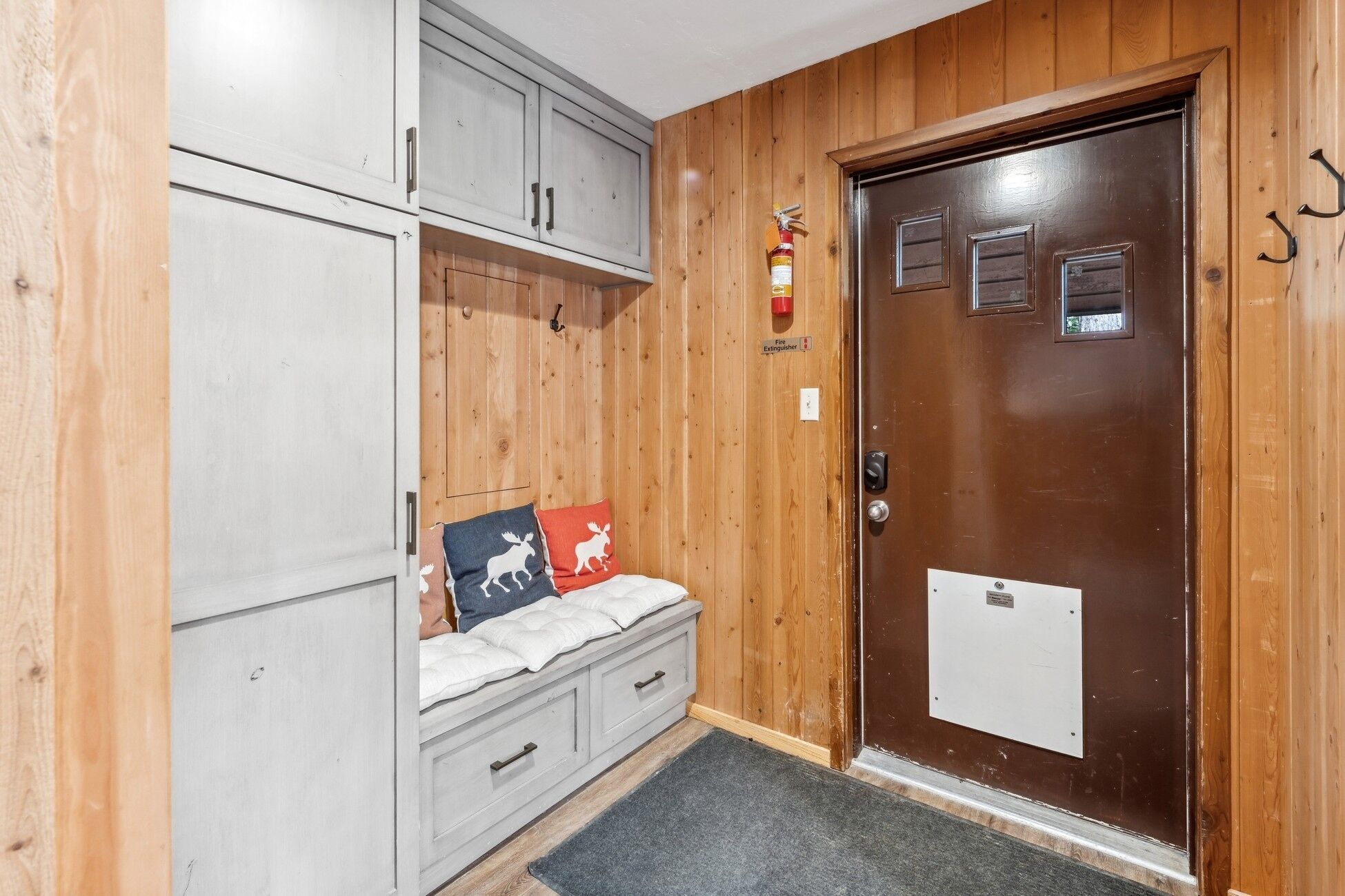 A room with a wooden door, a fire extinguisher, a bench with pillows, and a small cabinet.