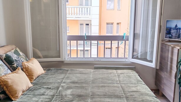Ein Schlafzimmer mit Bett, Fenster mit Blick auf Gebäude und einem Fernsehgerät auf einem Standfuß.
