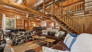 A log cabin interior with a dining area, a living room with a sofa and a coffee table, and a staircase. - Log Cabin Home w/ Private Hot Tub (Columbia Falls)