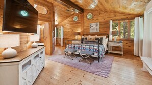 A cozy cabin interior with a wooden ceiling, a bed with a patterned bedspread, a desk with a lamp, and a large window with curtains. - Log Cabin Home w/ Private Hot Tub (Columbia Falls)
