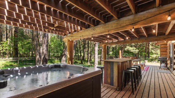 A wooden deck with a hot tub, bar area, and outdoor seating. - Log Cabin Home w/ Private Hot Tub (Columbia Falls)