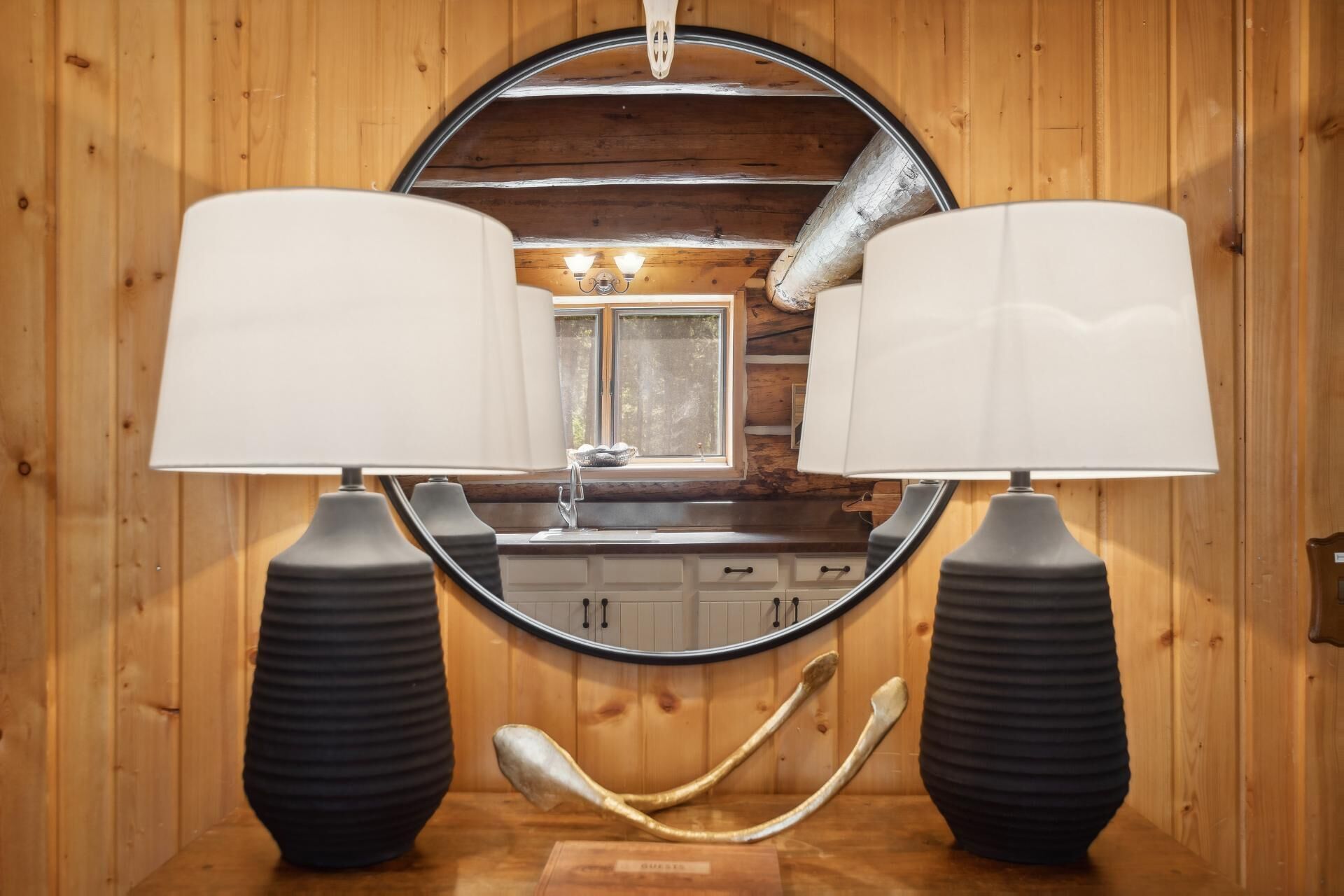 A wooden interior with a round mirror, two black lamps, and a sink with a window above.