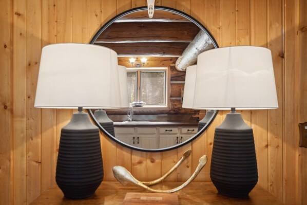 Un intérieur en bois avec un miroir rond, deux lampes noires et un lavabo placé sous une fenêtre.