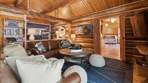 A cozy log cabin interior with a leather sofa, a round coffee table, and a bookshelf filled with books. - Log Cabin Home w/ Private Hot Tub (Columbia Falls)