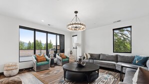 A spacious living room with a large window, a chandelier, and a modern sofa set. - Penthouse w/ Hot Tub, Fireplace and Firepit (Whitefish)