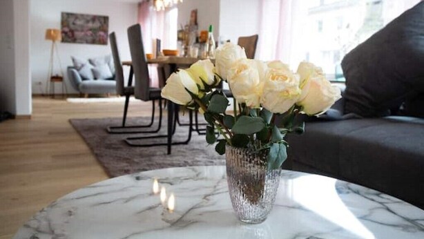 A living room with a marble coffee table, a vase with white flowers, a grey sofa, and a dining area in the background. - Traumhafte Stadtwohnung (Duisburg)