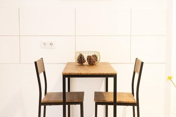 Un comedor sencillo con una mesa de madera y dos sillas, una pequeña cesta decorativa sobre la mesa, y una pared de azulejos blancos con un enchufe.