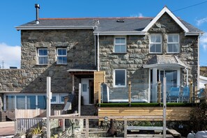A stone house with a wooden deck, a covered porch, and a small garden area.