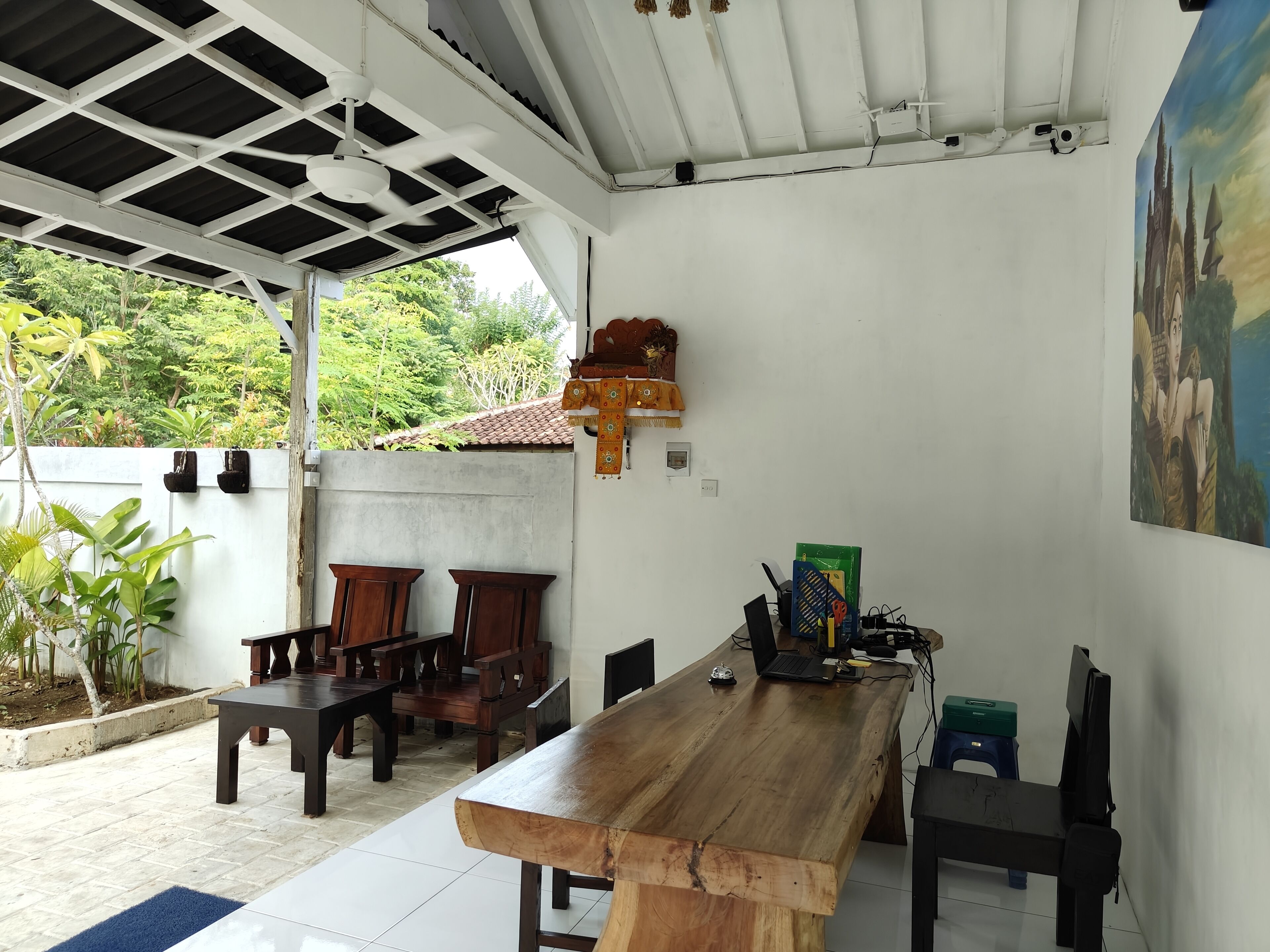 A covered outdoor dining area with a wooden table and chairs, a laptop, and a painting on the wall.