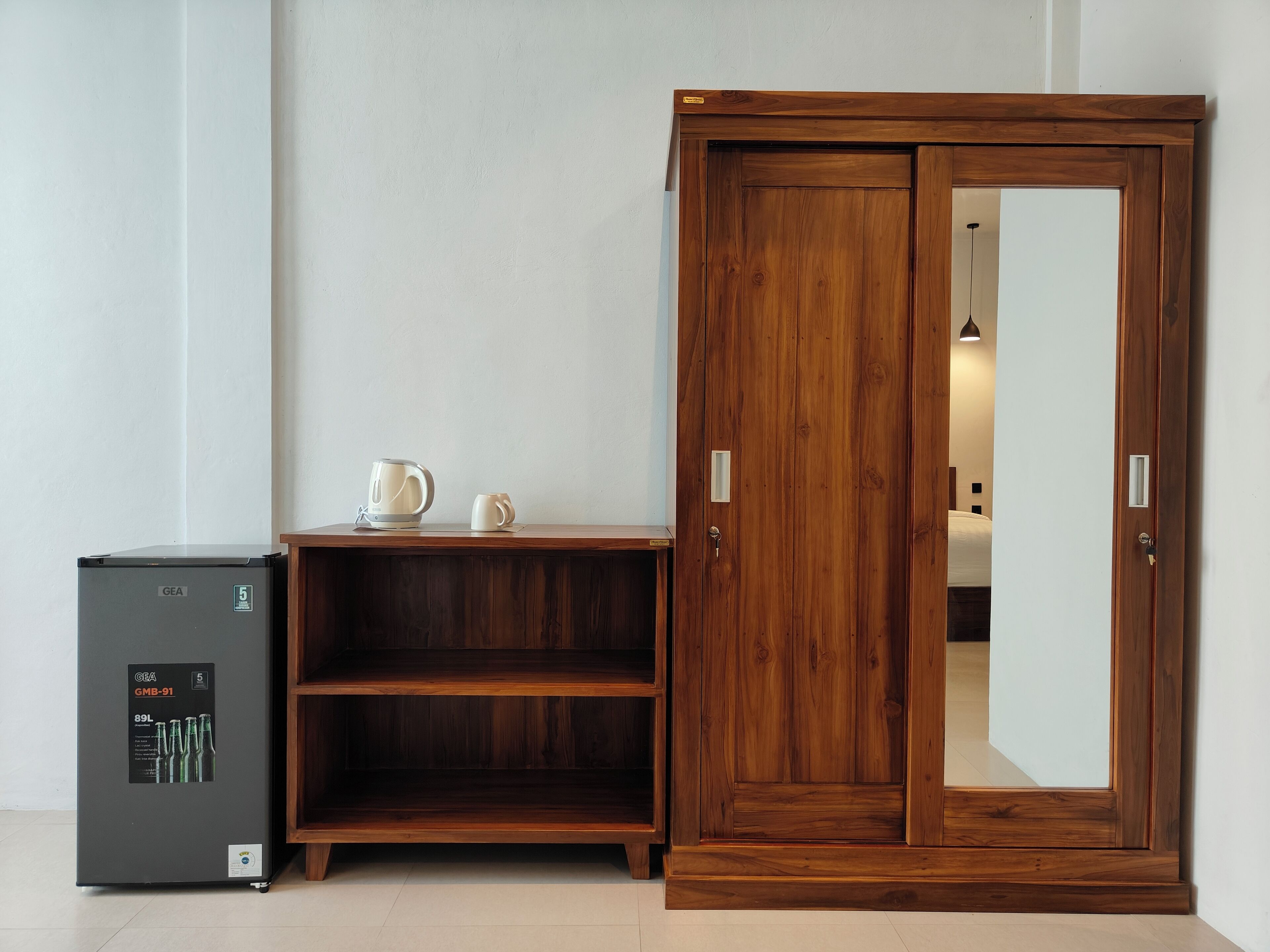 A compact room with a wooden wardrobe, a small table with a kettle and mug, and a GEA refrigerator.