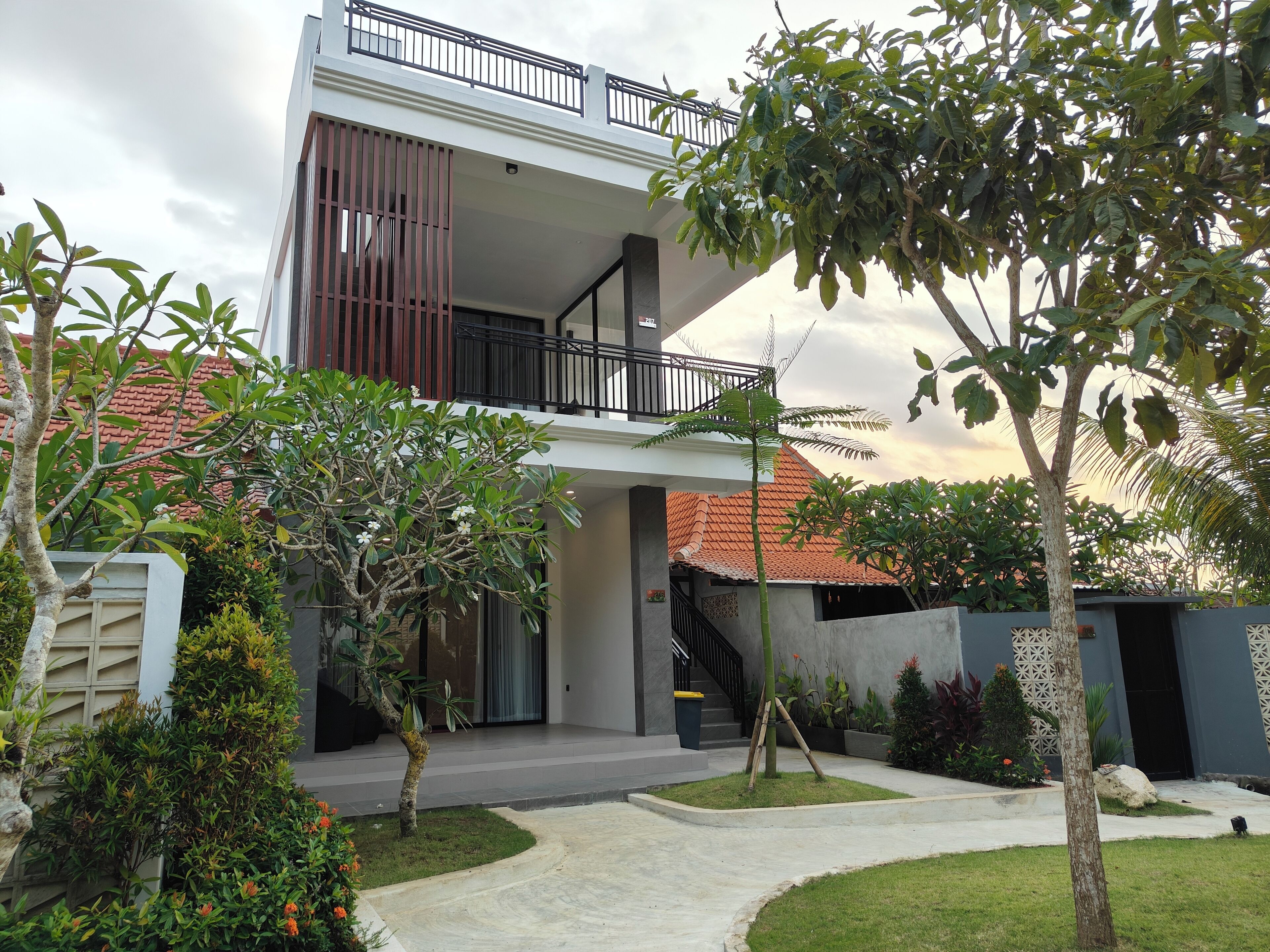 A modern two-story house with a balcony, surrounded by well-maintained gardens and a clear sky.