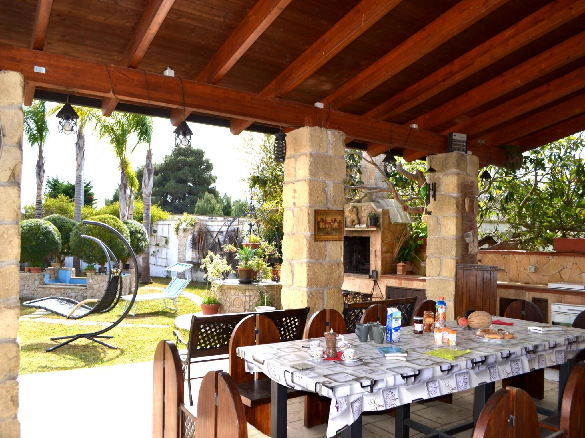 Un patio al aire libre con una mesa lista para comer, rodeado de pilares de piedra, y vistas a un jardín con árboles y una piscina.