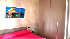 Un dormitorio con cama, armario de madera, un cuadro colgado en la pared que muestra una escena costera y una mesita de noche con lámpara.