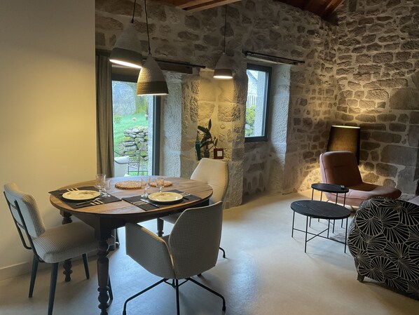 A dining area with a stone wall, a wooden table set for four, and a view of greenery outside. - Gîte du Four à Pain, Domaine de la Voute, Plateau de Millevaches (Viam)
