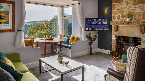 A cozy living room with a stone fireplace, a television mounted on the wall, and a view of the outdoors through large windows.