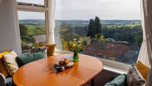 A round wooden table with a croissant and a vase of flowers, set against a window overlooking a rural landscape.