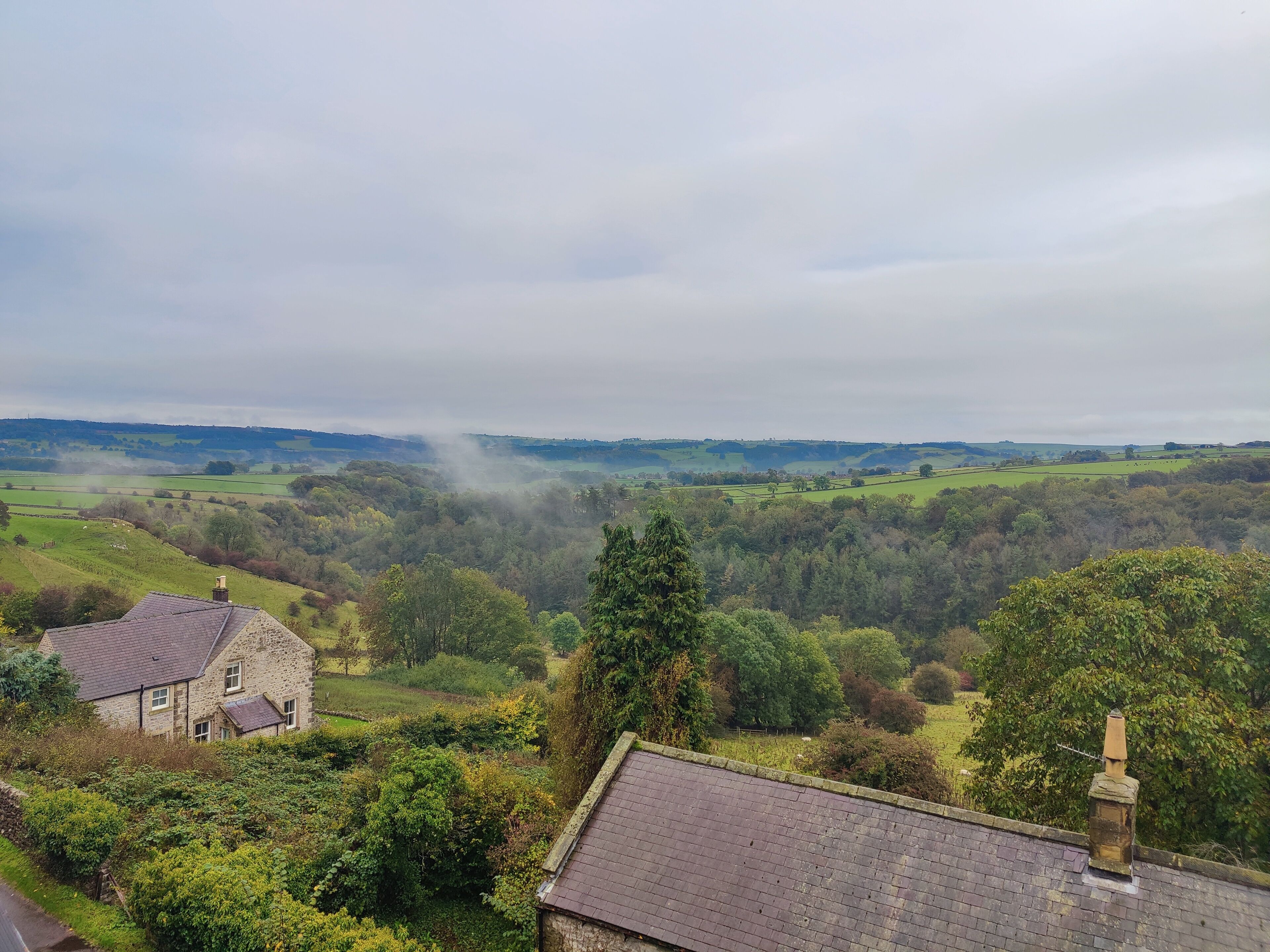 Une maison en pierre, dotée d’un toit en pente et d’une cheminée, entourée d’une végétation luxuriante et de champs.