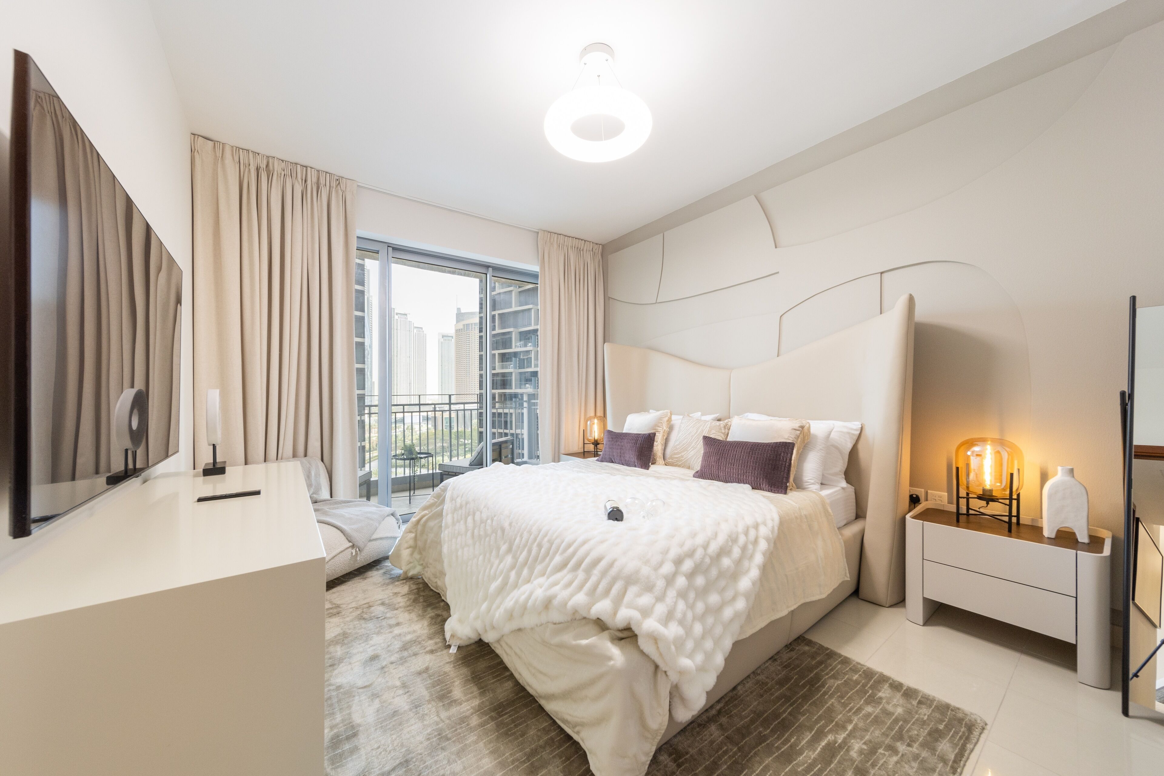 A modern bedroom with a large bed, a bedside table with a lamp, a mirror, and a view of the city through the window.