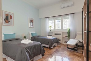 Habitación de hotel con dos camas, una ventana con cortinas, un radiador, una mesita con lámpara y una silla.