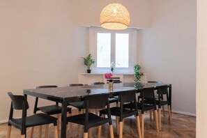 Un coin repas avec une table rectangulaire, des chaises noires, un luminaire suspendu en fibres tressées et des plantes en pot disposées sur une étagère blanche.