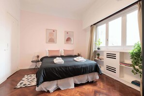 Une chambre à coucher comprenant un lit, des tables de chevet, une fenêtre avec des rideaux, une plante en pot et une étagère fixée au mur.