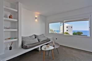 Un salon moderne avec un canapé, une petite table, des étagères ornées d’objets décoratifs et une grande fenêtre offrant une vue sur la mer et les bâtiments.