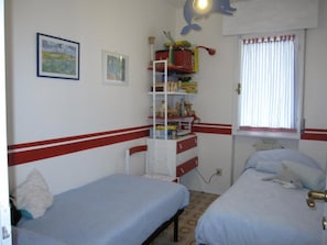 A room with two beds, a shelf unit with various items, a window with curtains, and a wall-mounted light fixture.