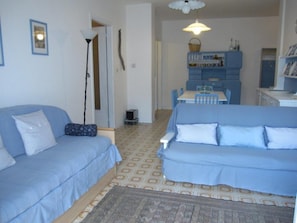Un salon avec deux canapés bleus, un tapis à motifs, un coin repas avec une table et des chaises, et une kitchenette avec des meubles de couleur bleue.