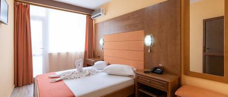 A hotel room with a bed, a bedside table with a phone, a mirror, and a window with curtains.