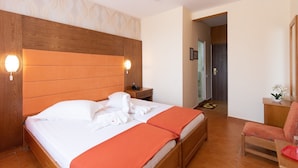 A hotel room with a wooden headboard, a bed with white linens and an orange throw, a wooden nightstand with a phone, and a small table with a vase of flowers. - HOTEL DELTA (Constanta)