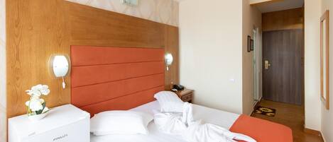 A hotel room with a wooden headboard, a white bed, a nightstand with a phone and a lamp, and a small table with a vase of flowers.