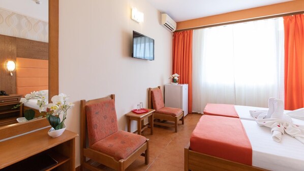Habitación de hotel con dos camas individuales, un escritorio con silla, una mesita con otra silla, un televisor montado en la pared y una ventana con cortinas naranjas.