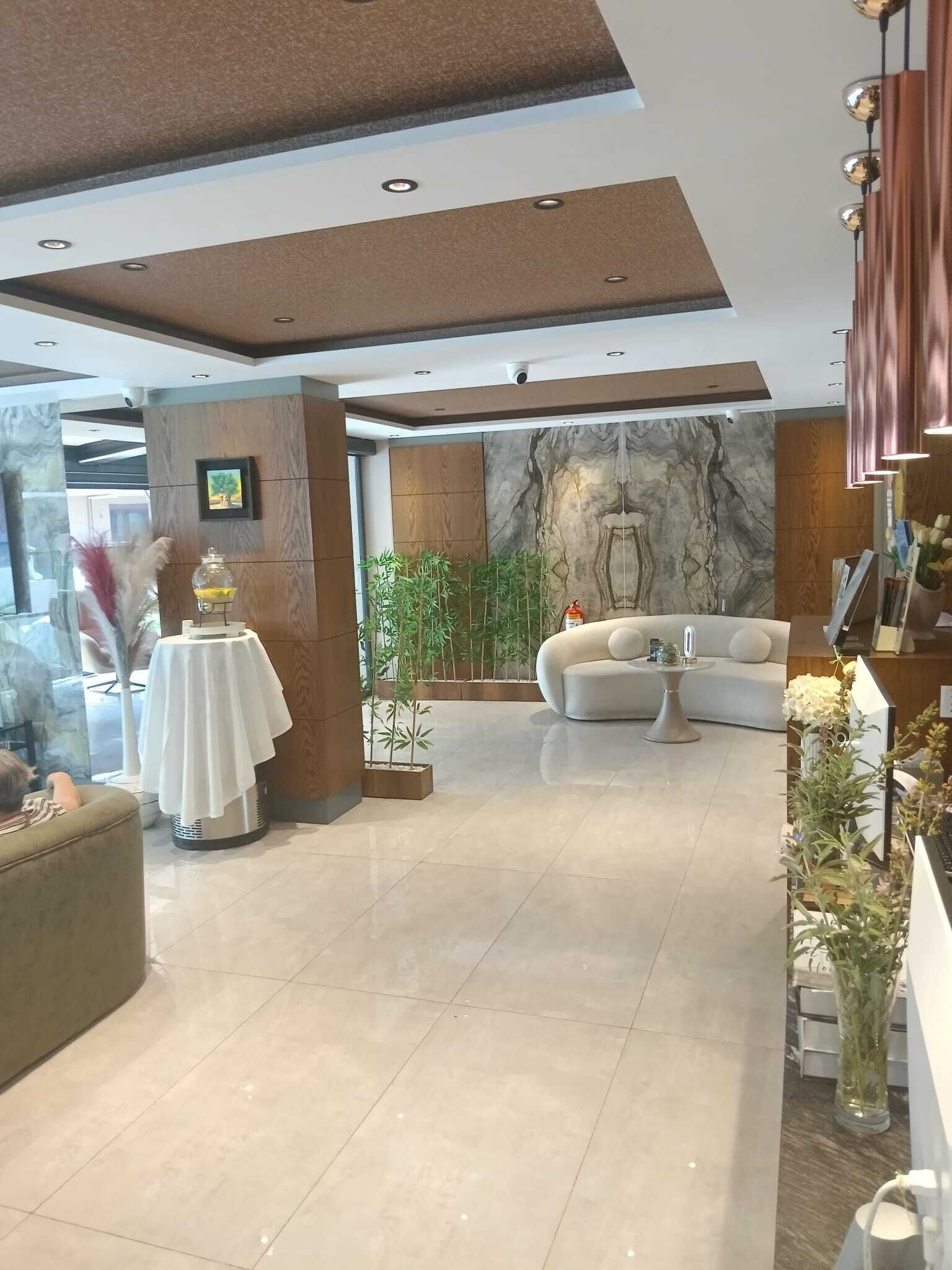 A modern hotel lobby with a sofa, a table with a white tablecloth, a wall with a marble-like design, and a large window.