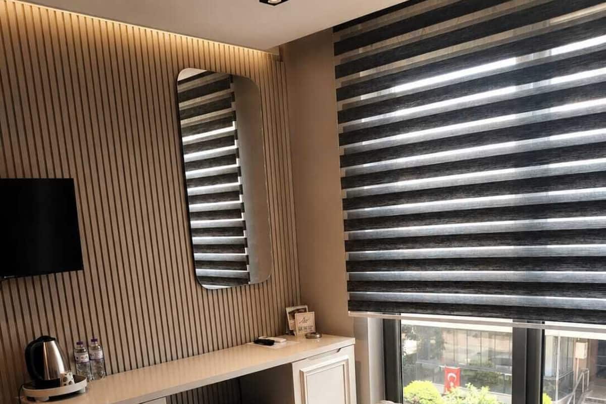 A modern hotel room with a bed, a desk, a mirror, and a window with blinds.