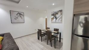 Un salon moderne avec un canapé gris, une table à manger avec quatre chaises et un réfrigérateur. Deux œuvres d’art encadrées ornent les murs.