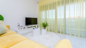 Una sala moderna con un sofá, un televisor de pantalla plana, dos mesitas blancas, una planta en maceta y cortinas translúcidas amarillas.
