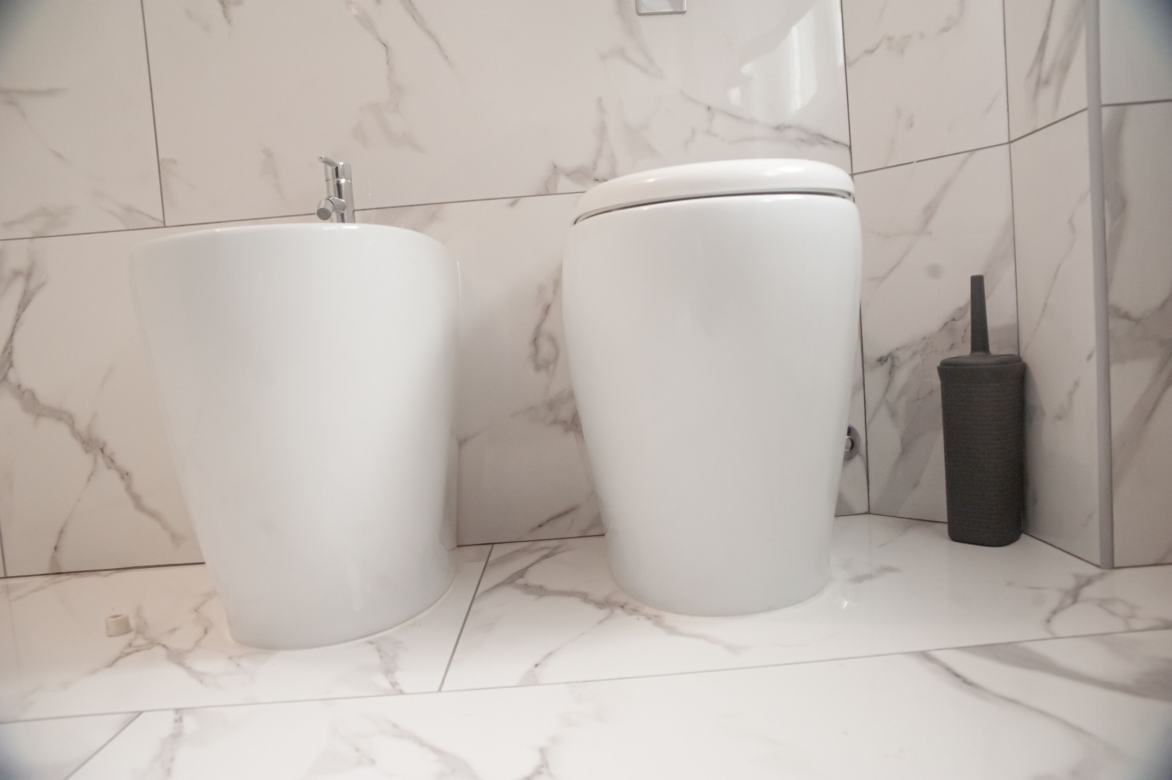 Un baño con inodoro y lavamanos blancos, baldosas de mármol y un escobillón negro.