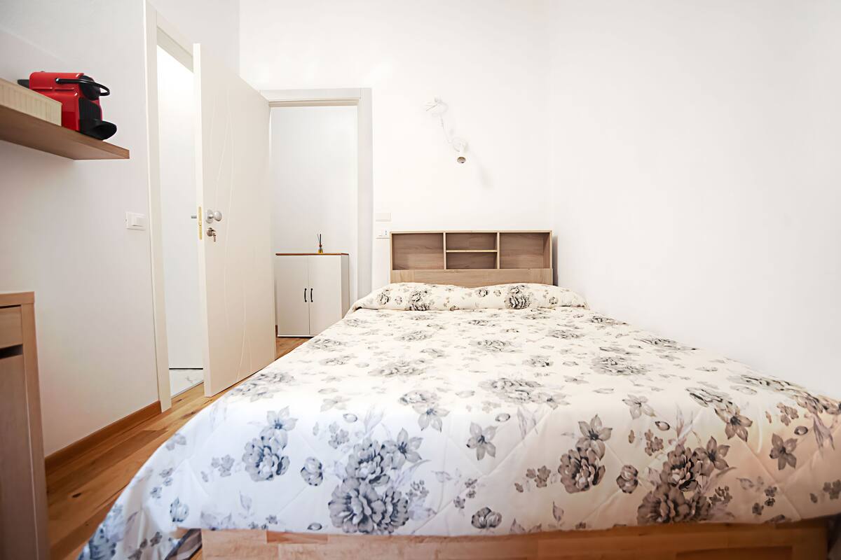 A bedroom with a floral bedspread, a wooden headboard, and a shelf with a red object.