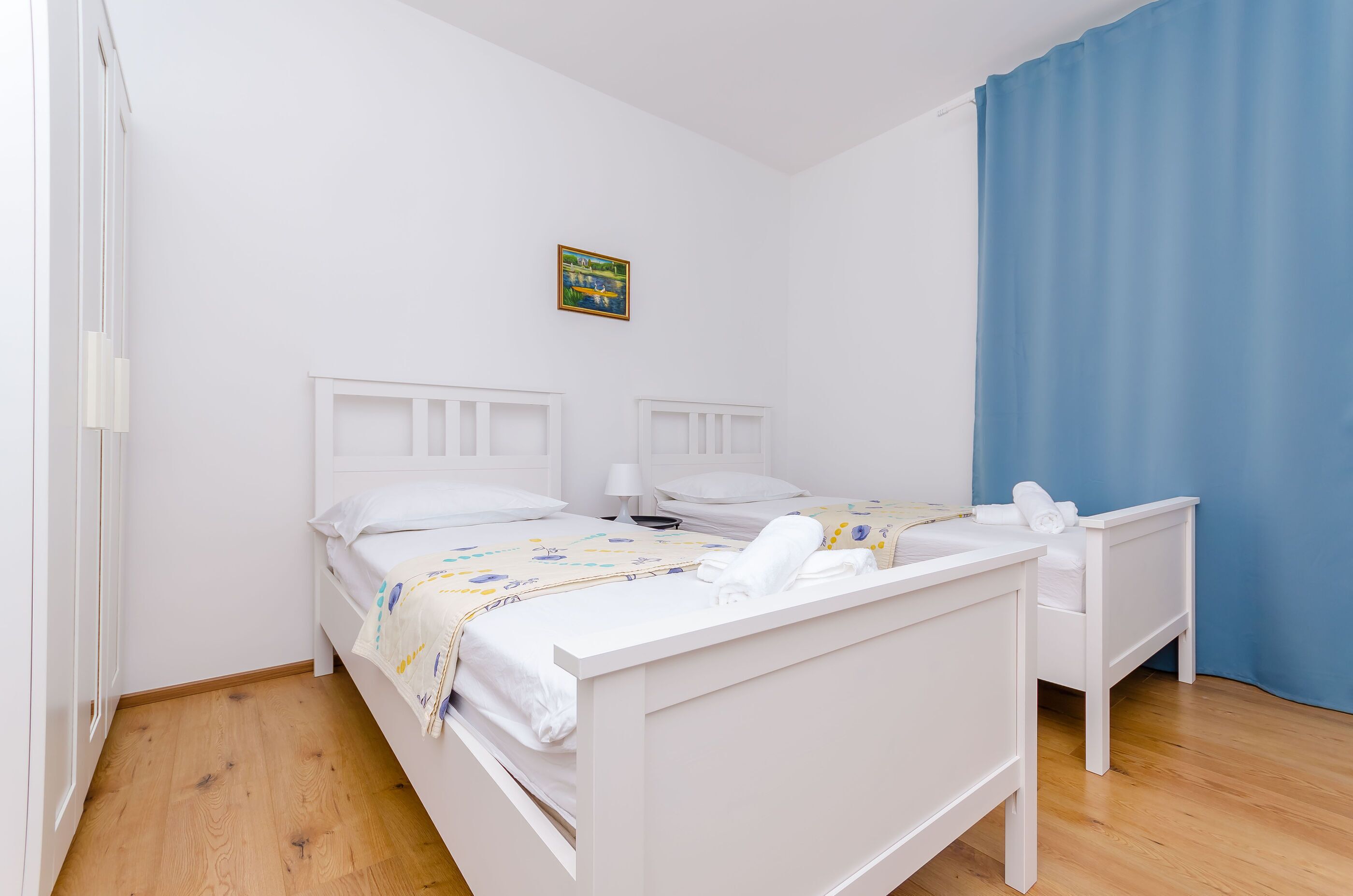 A clean, white bedroom with two beds, a small bedside table with a lamp, and a blue curtain.
