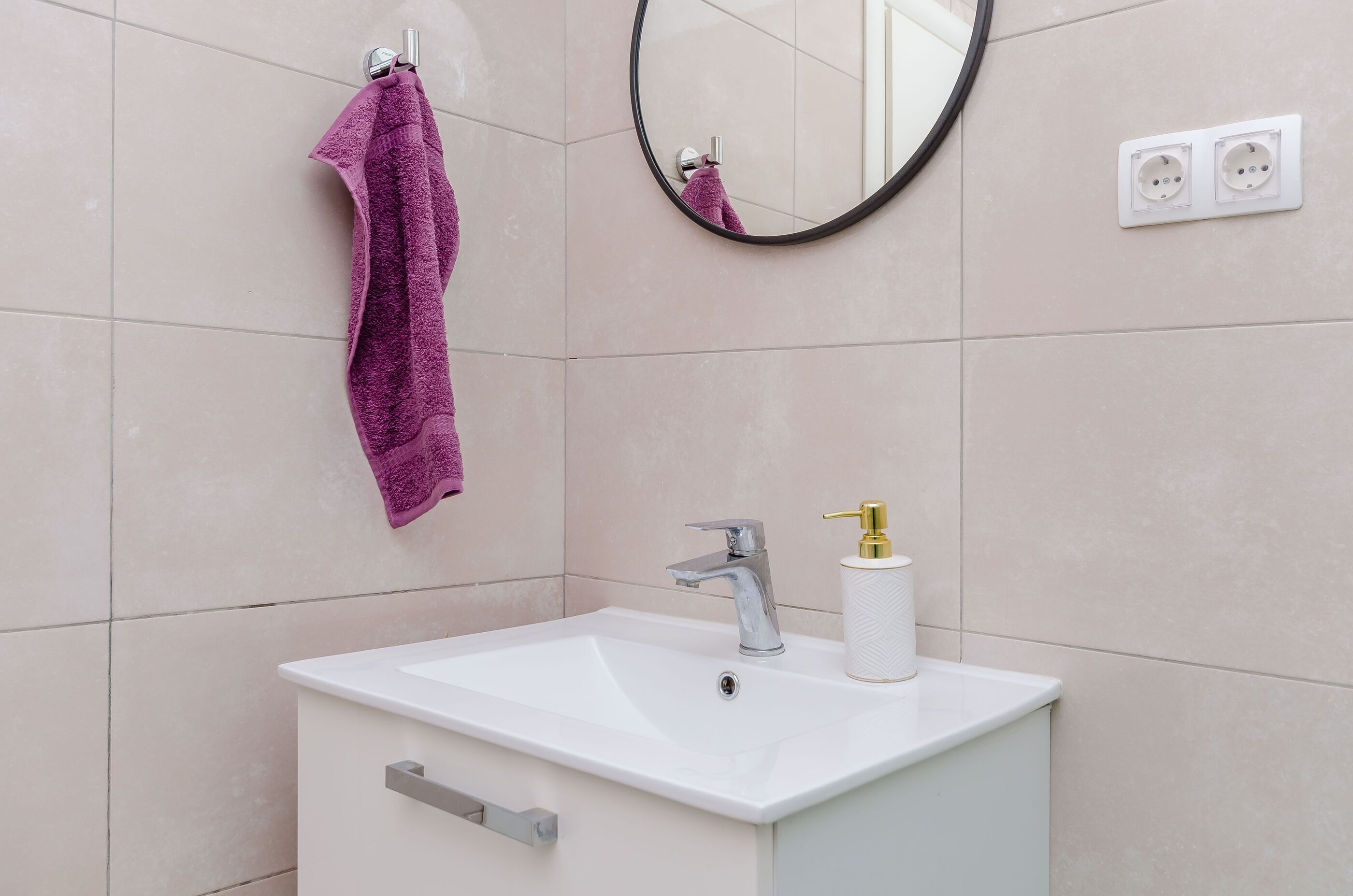 Une salle de bain avec un lavabo blanc, une serviette violette, un miroir et une prise électrique au mur.