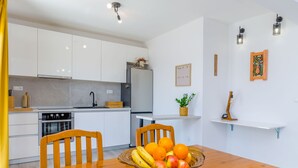 A modern kitchen with white cabinets, a refrigerator, and a dining table with a fruit bowl. - Apartment Rizza (Trogir)
