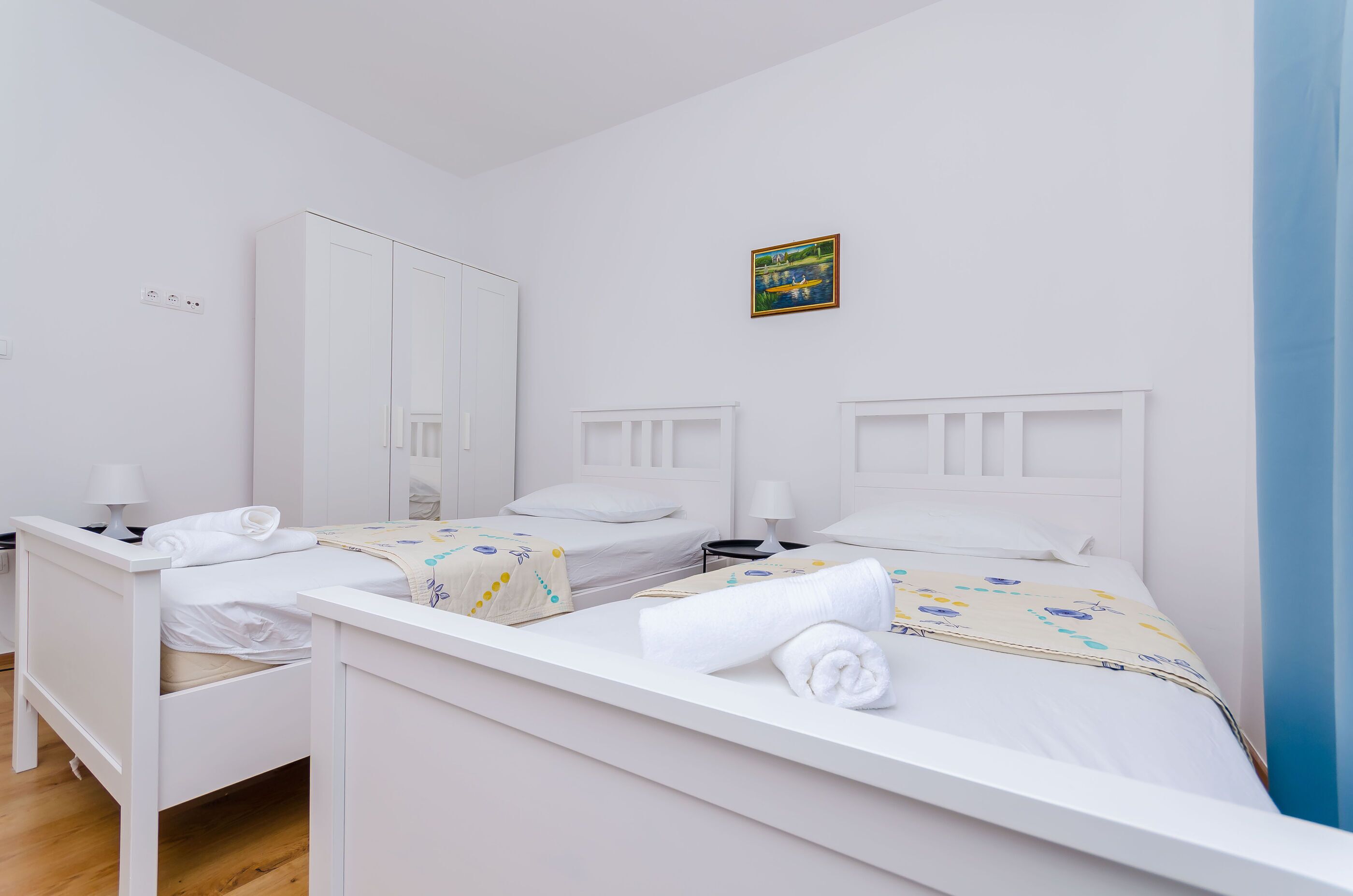A twin room with two beds, each with a white headboard, a bedside table, and a lamp. There is a wardrobe and a painting on the wall.