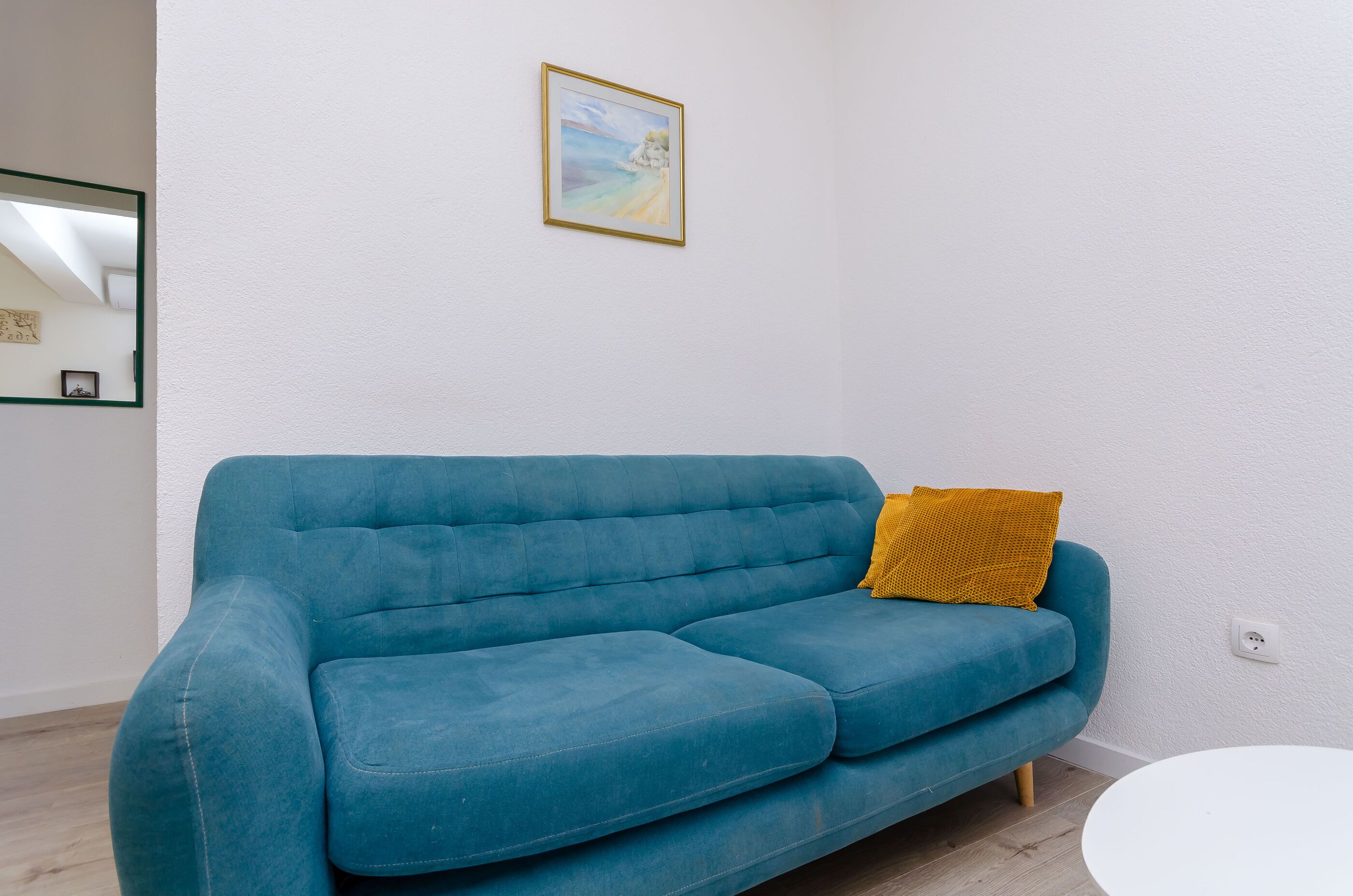 Ein blaues Sofa mit einem senfgelben Kissen in einem Raum mit weißen Wänden und einem darüber hängenden Bild.