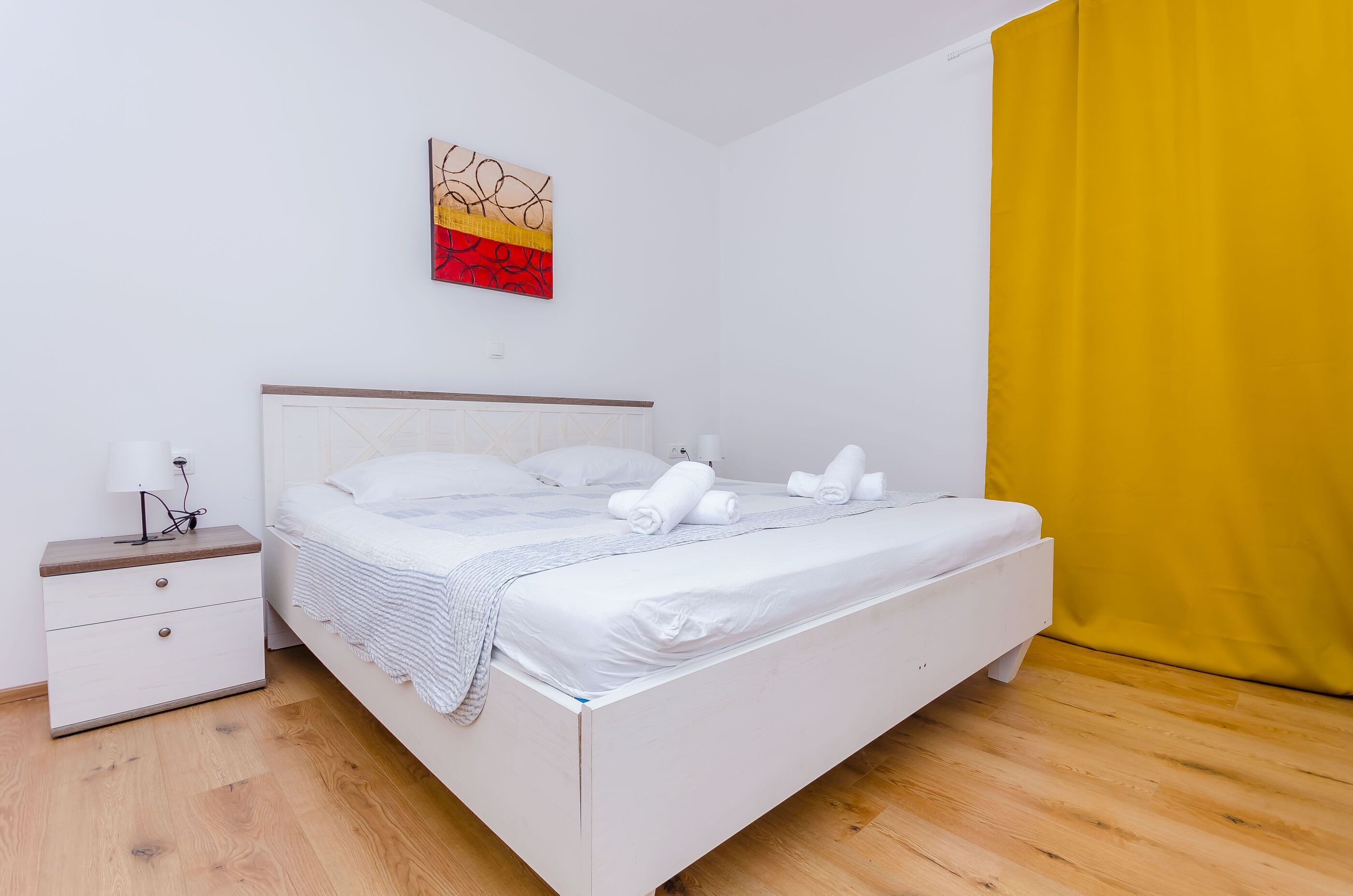 A neatly made bed with white linens and rolled towels, a bedside table with a lamp, and a wall art piece above the bed.