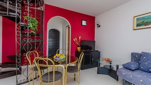 A modern living room with a red accent wall, a staircase with a wrought iron railing, a dining area with a glass table and chairs, and a blue sofa. - House Zdenka (Okrug Donji)
