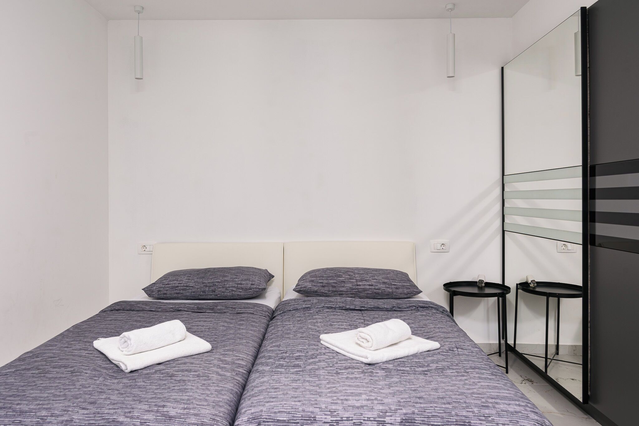 A twin bed with grey bedding and two white towels, a small bedside table, and a mirror on the wall.
