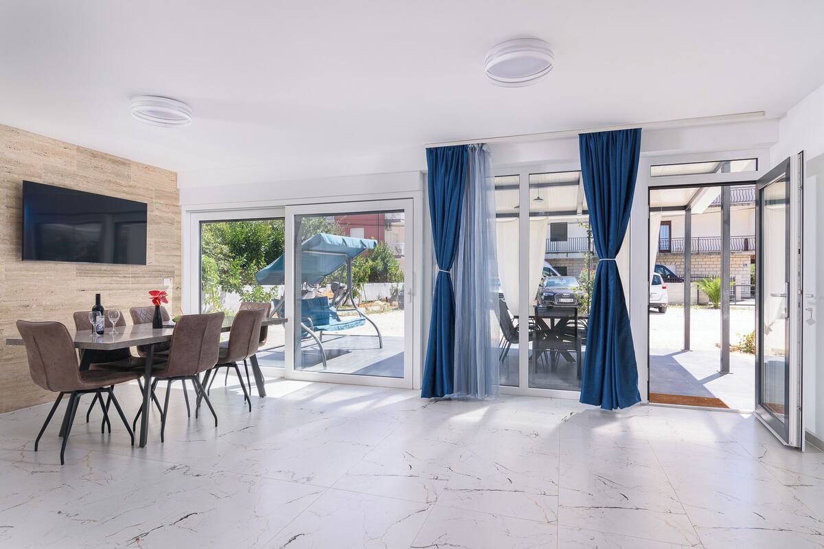 A dining area with a table and chairs, a flat-screen TV mounted on a wooden wall, and a view of the outdoors through large glass doors.
