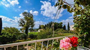A balcony with a white railing, a table with a vase of flowers, and a view of a garden and distant buildings. - Apartments Helios (Kastel Gomilica)