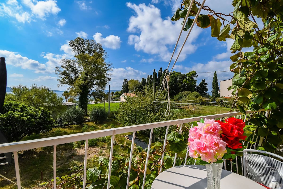 A balcony with a white railing, a table with a vase of flowers, and a view of a garden and distant buildings.