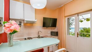 A kitchen with a glass table, red chairs, a TV, and a view of greenery through the door. - Apartments Helios (Kastel Gomilica)